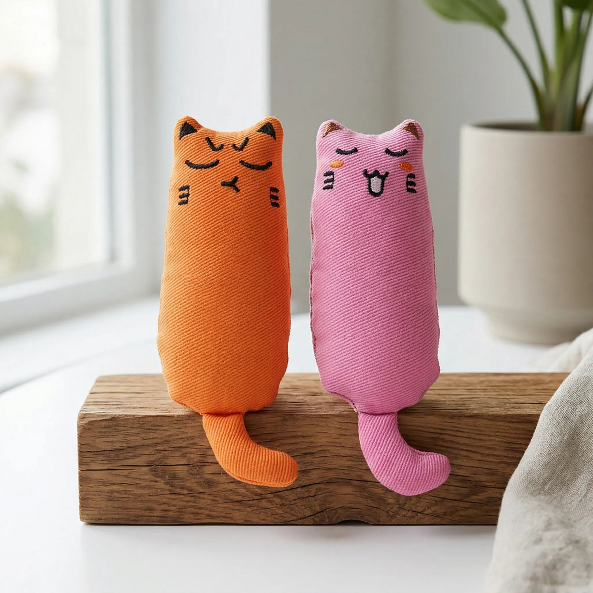 Deux jouets en peluche pour chat avec herbe à chat intégrée, un orange et un rose, posés sur un bloc en bois décoratif.
