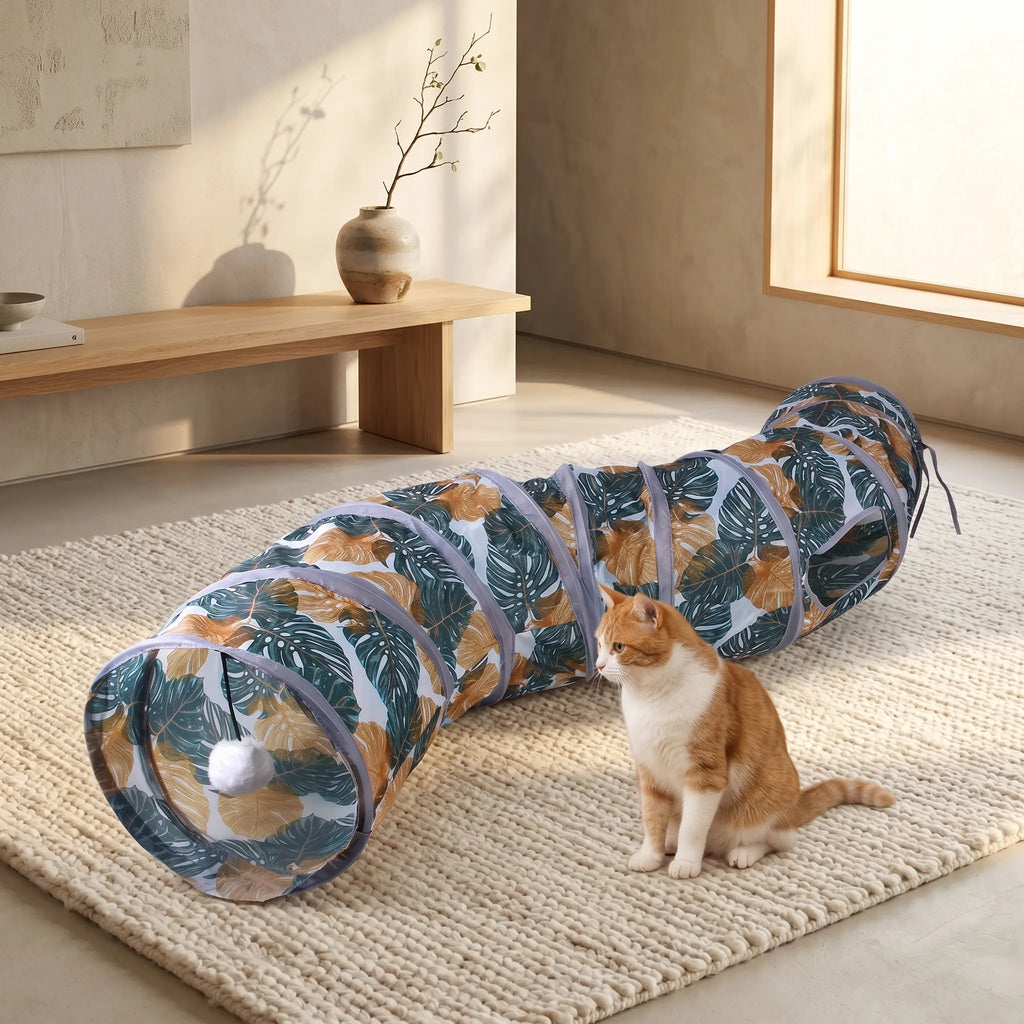 Tunnel pour chat pliable avec motif de feuilles de monstera sur un tapis beige, à côté d'un chat roux assis.