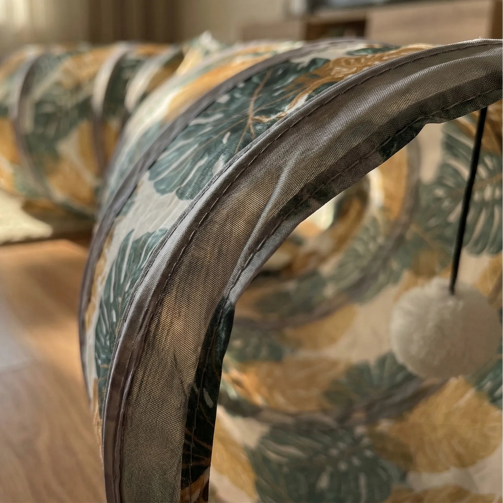 Gros plan sur l'entrée d'un tunnel pour chat montrant le tissu à motif tropical, l'armature métallique et un jouet pompon suspendu