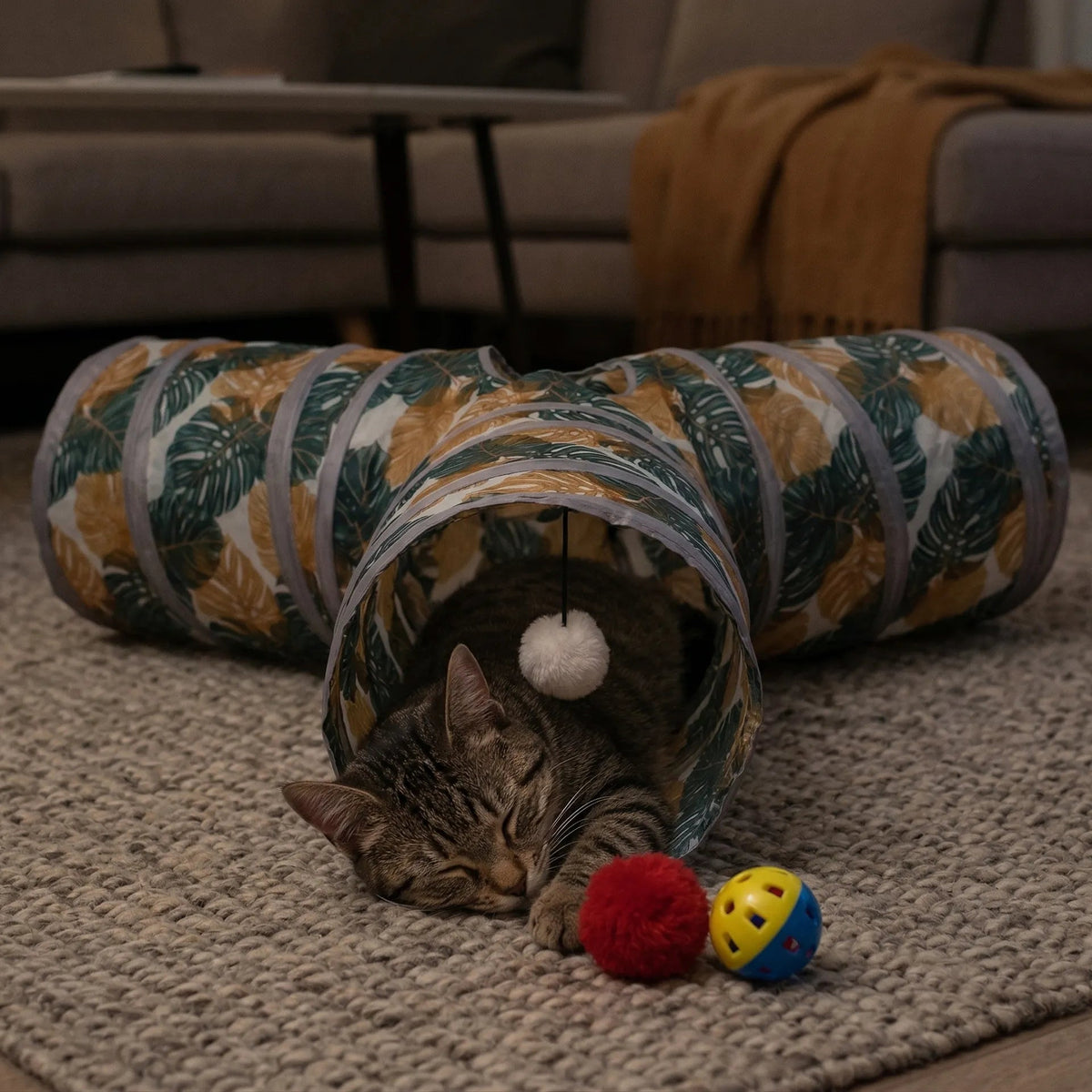Chat tigré dormant paisiblement à l'intérieur d'un tunnel de jeu à motif végétal, à côté de balles colorées pour chat